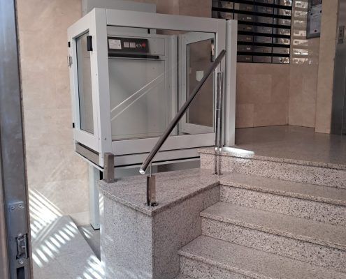 "Resultado final: Instalación de plataforma elevadora vertical de corto recorrido con cerramiento acristalado. Una solución segura, moderna y perfectamente integrada con la estética de granito y mármol del edificio, garantizando la total accesibilidad desde la entrada." Plataforma elevadora vertical instalada junto a la escalera de granito en un portal de Murcia.